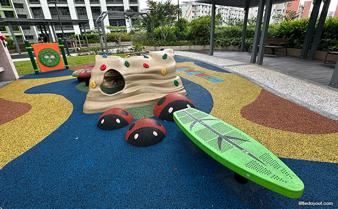 A rock-shaped climbing tunnel, leaf-shaped balance feature and red ladybird obstacles on blue surfacing.