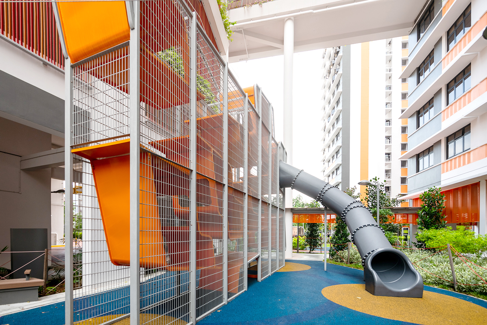 Orange climbing wall enclosed in metal mesh, with a grey tube slide, at the Kallang Residences King Crawler playground.