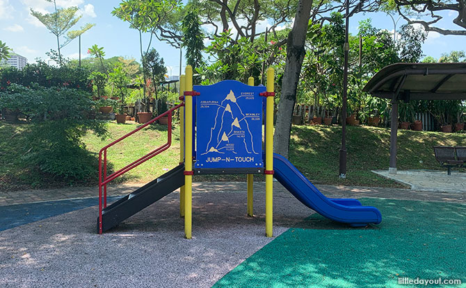 A low blue toddler slide with yellow posts and nearby blue activity panels at Aida Park.